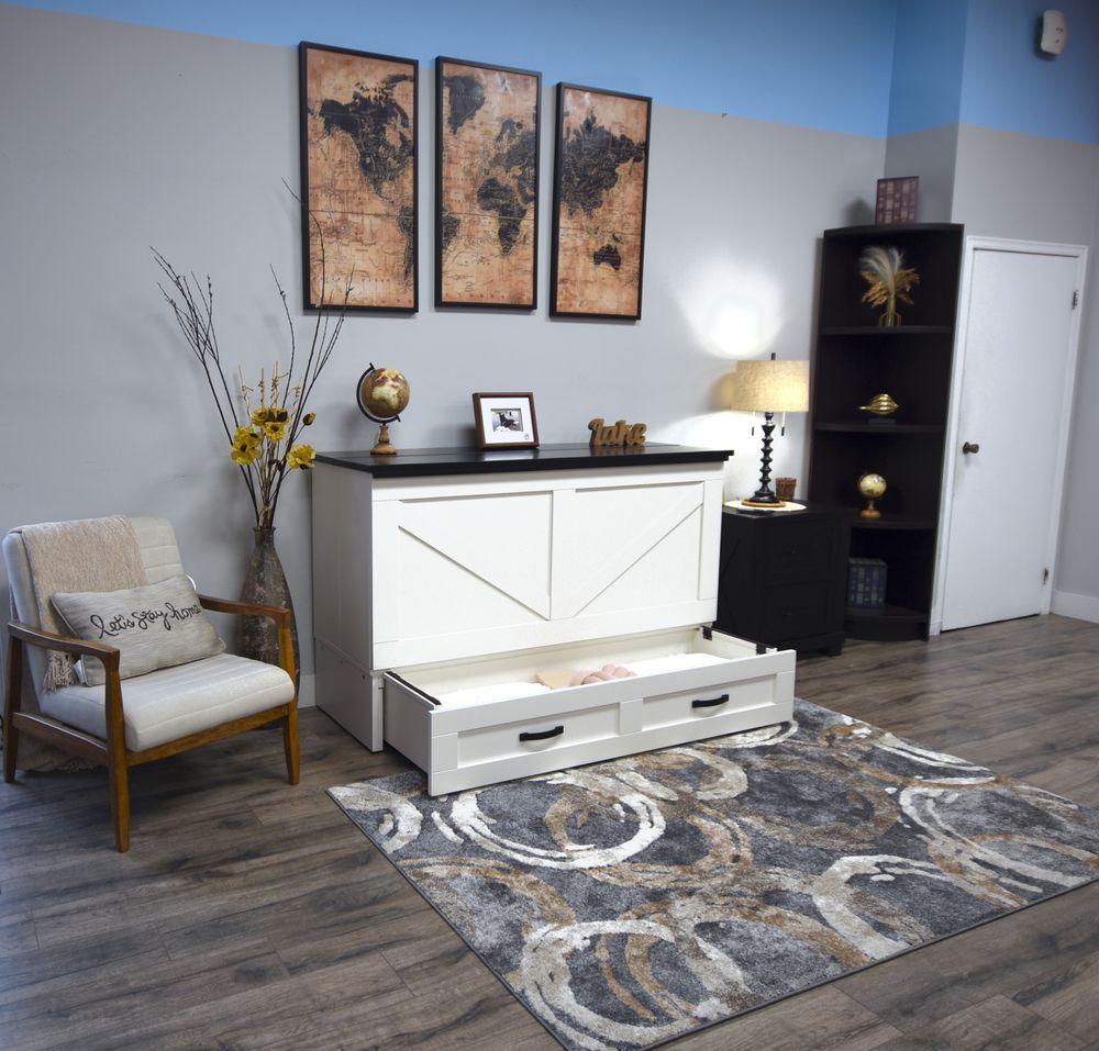 A white storage chest with a pull-out drawer underneath, shown in a styled room setting with a chair, rug, shelving, and wall art.
