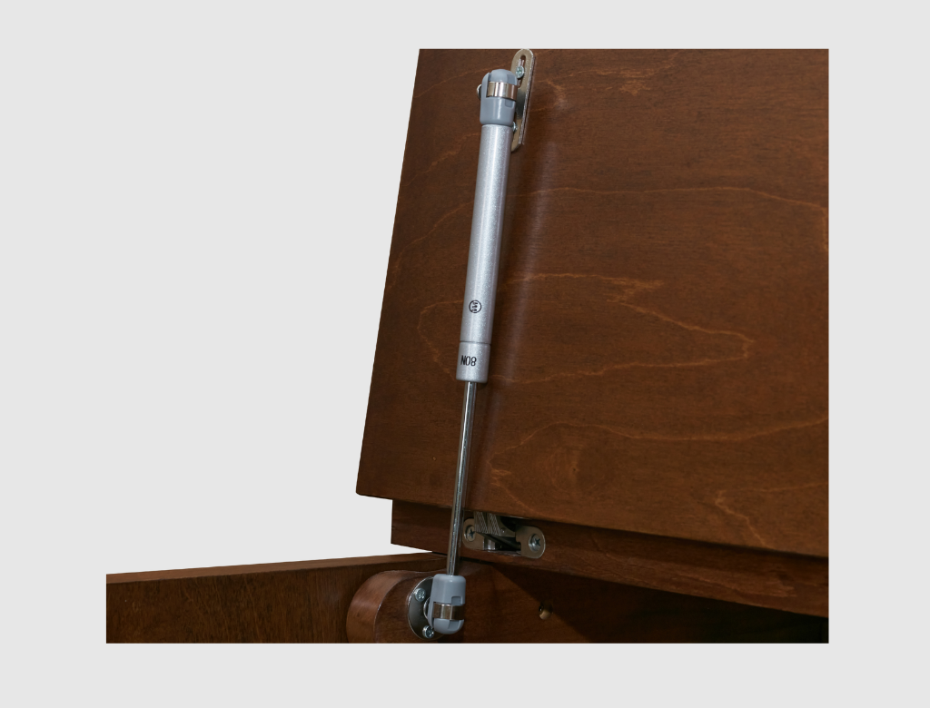 A silver metal cabinet hydraulic lift handle shown in close-up, mounted on a brown wooden cabinet door with the lower cabinet panel slightly visible below.